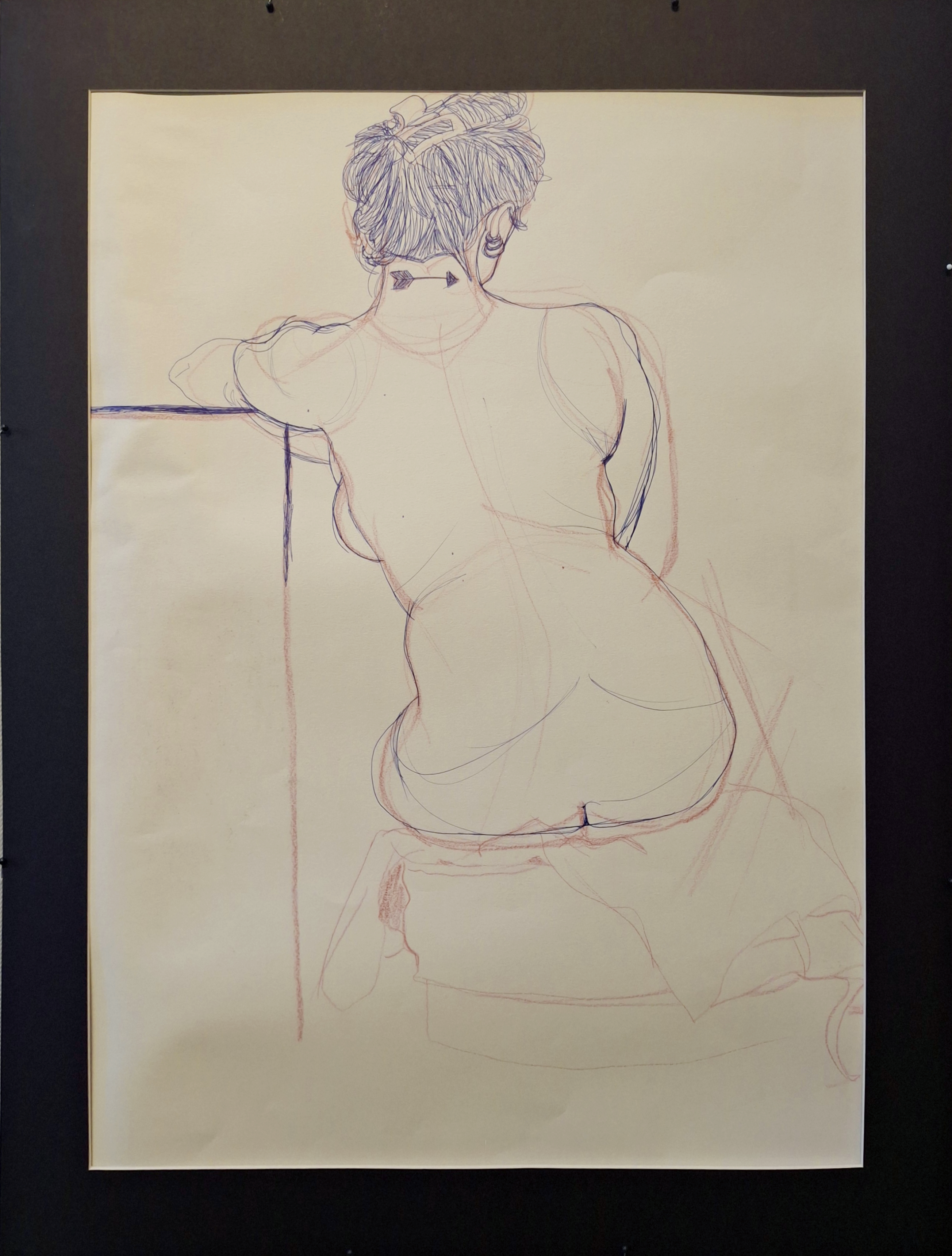 
A photograph of a figure drawing sitting with the model's back facing the viewer. The piece was done in red colored pencil as an undersketch and finalized with a blue pen. The model has their hair in a bun with a clip and their left arm resting on a podium. The model is also sitting on a stool with blankets to cover the seat.